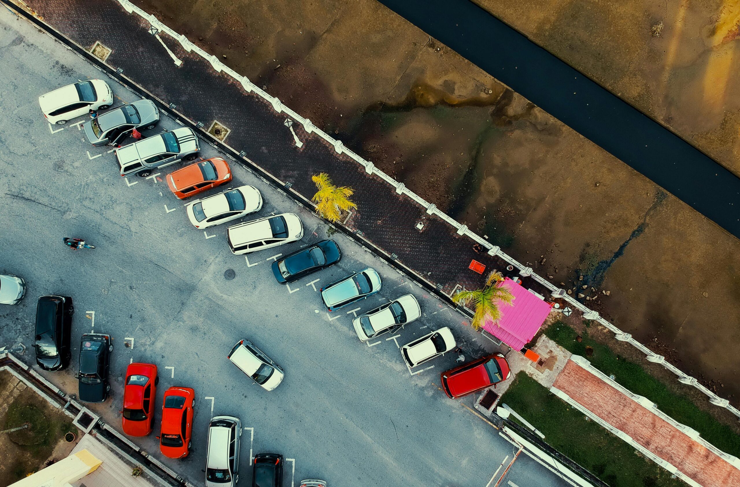 Aerial shot of a parking lot with cars and a pink building, next to a waterway.